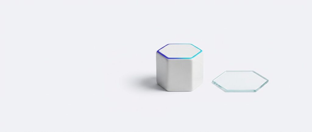 A white ceramic hexagonal seal stamp standing upright beside a thin clear glass hexagon plate, lit by a soft indigo to cyan edge light, on a flat light grey background.